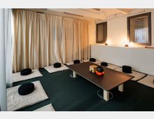 Ein schlichter Meditationsraum mit einem niedrigen Tisch in der Mitte, darauf liegen eine Klangschale und ein kleiner Korb. Um den Tisch sind auf dem Teppichboden weiße Matten mit schwarzen Meditationskissen angeordnet. Vorhänge bedecken die Wände, und zwei Fenster mit indirekter Beleuchtung befinden sich an der rechten Wand.