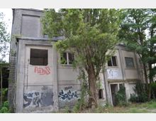 Verlassenes zweistöckiges Betongebäude mit beschädigten Fenstern und Wänden, bedeckt mit Graffiti. Umgeben von überwucherter Vegetation und Bäumen, die teilweise die Fassade verdecken.