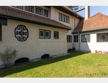 Zweistöckiges Haus mit hellen verputzten Wänden, rot gedecktem Satteldach und mehreren Fenstern mit kunstvoll verzierten Gitterrahmen. Im Vordergrund ein gepflegter Rasen, umgeben von vereinzelten Pflanzen.