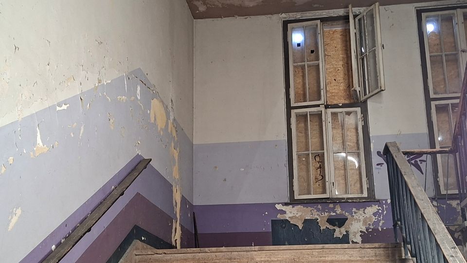 Abgenutztes Treppenhaus in einem älteren Gebäude mit stark abblätternder Farbe an den Wänden und mehrfarbigen, diagonalen Streifen entlang des Treppenlaufs. Hohe Fenster mit teils geöffneten Rahmen und teils verbretterten Scheiben lassen Licht in den Raum; ein Metallgeländer führt nach oben.