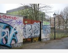 Eine mit Graffiti bedeckte Mauer und ein Tor aus Metall, das zu einem eingezäunten Bereich führt. Im Hintergrund sind Bäume ohne Laub, ein modernes Gebäude mit grüner Fassade und ein älteres Backsteingebäude zu sehen.