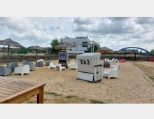 Ein Sandbereich mit Strandkörben, weißen und grauen Stühlen sowie kleinen Strohpavillons. Im Hintergrund sind eine Steinmauer, ein modernes Gebäude und eine blaue Bogenbrücke zu sehen.