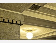 Ansicht einer Decke mit texturierter Oberfläche und einem dekorativen rechteckigen Mosaik. Eine hängende Deckenleuchte mit runder Glühbirne und sternförmigem Ornament befindet sich in der Mitte; ein Pfeiler mit geriffelten Steinmustern ist im Vordergrund sichtbar.