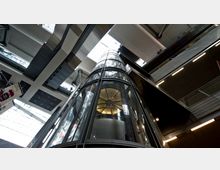 Blick nach oben in das Innere eines modernen Gebäudes mit einem gläsernen Aufzug in der Mitte. Die Umgebung zeigt mehrere Ebenen mit metallischen und gläsernen Elementen sowie Sicht auf ein Dachfenster, durch das Tageslicht einfällt.