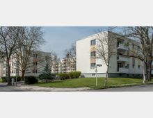 Mehrstöckige Wohngebäude in einem Wohngebiet mit hellen Fassaden und Balkonen, umgeben von Bäumen, Sträuchern und gepflegten Grünflächen. Im Vordergrund sind Gehwege, Straßenlaternen und ein Straßenschild zu sehen.