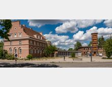 Vorstadtlandschaft mit einem rosafarbenen Gebäude im klassizistischen Stil mit weißen Fensterbögen links und einem Backsteingebäude mit einem Turm rechts. Im Vordergrund ein gepflasterter Platz mit Blumenbeeten, Sitzbänken und Straßenschildern, umgeben von Bäumen und unter einem blauen Himmel mit Wolken.