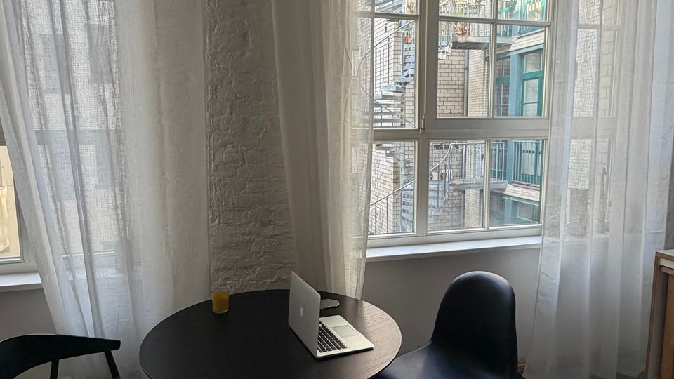 Heller Innenraum mit großen Sprossenfenstern und weißen, halbtransparenten Vorhängen an einer freigelegten Ziegelwand. Vor den Fenstern steht ein runder Tisch mit Stühlen und Hocker; durch die Fenster sind ein Innenhof mit Backsteinfassaden und eine Außentreppe zu sehen.