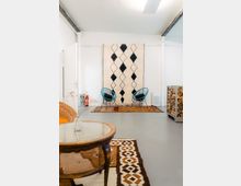 Minimalistisch eingerichteter Raum mit hellen Wänden, dekoriert mit einem großen beige-schwarzen Teppich an der Wand. Im Vordergrund ein runder Holztisch mit Glastischplatte, im Hintergrund zwei blaue Stühle auf bunten Teppichen.