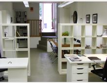 Modernes Büro mit weißer Einrichtung, Trennregalen und Schreibtischen. Im Hintergrund befinden sich ein Konferenztisch mit Stühlen, ein Dartscheibe an der Wand sowie eine offene Tür, die zu einem erhöhten Außenbereich führt.