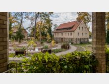 Ein historisches Gebäude mit cremefarbener Fassade und rotem Ziegeldach steht inmitten eines gepflegten Gartens mit gewundenen Wegen, dekorativen Pflanzenbeeten und Schildern. Im Vordergrund ist eine überdachte Pergola mit rankenden Pflanzen sichtbar.