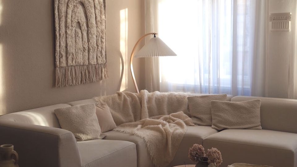 Sunlit living room with a cream sectional sofa, textured throw pillows and blankets, and a low round wooden coffee table on a shag rug. A large window with sheer curtains, a curved floor lamp, and a woven wall hanging decorate the neutral-toned space with wood flooring.