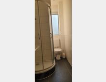 Kleines Badezimmer mit Eckdusche mit Glaswänden, einer Toilette und einem Fenster mit Milchglas. Die Wände sind weiß gefliest, der Boden besteht aus dunkelgrauen Fliesen.