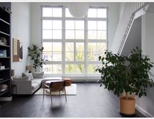 Wohnzimmer mit hohen Decken und großer Fensterfront, die viel Tageslicht hereinlässt. Der Raum ist modern eingerichtet mit einem Sofa, einem Sessel, Pflanzen in Töpfen, einer schwarzen Bücherwand und einer offenen Treppe an der rechten Wand.