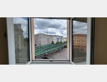 Blick aus einem geöffneten Fenster auf eine städtische Straße mit einer grünen Hochbahnbrücke und mehreren mehrstöckigen Wohn- und Geschäftshäusern. Im Hintergrund ist ein bewölkter Himmel über den Gebäudefassaden zu sehen.