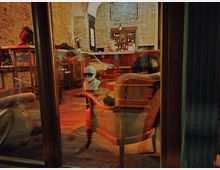 Innenansicht eines gemütlichen Cafés oder Bars mit Backsteinwänden, Holzstühlen und Tischen. Im Vordergrund steht eine Schaufensterpuppe mit Hut, dahinter eine Theke mit Flaschen und Dekoration.