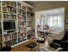 Wohnzimmer mit einem großen Bücherregal, das die linke Wand bedeckt, und einem Fernseher in das Regal integriert. Im angrenzenden Bereich befindet sich ein heller Raum mit großem Fenster, weißen Vorhängen, einer weißen Couch, mehreren Stühlen und dekorativen Elementen wie Bildern und Lampen.