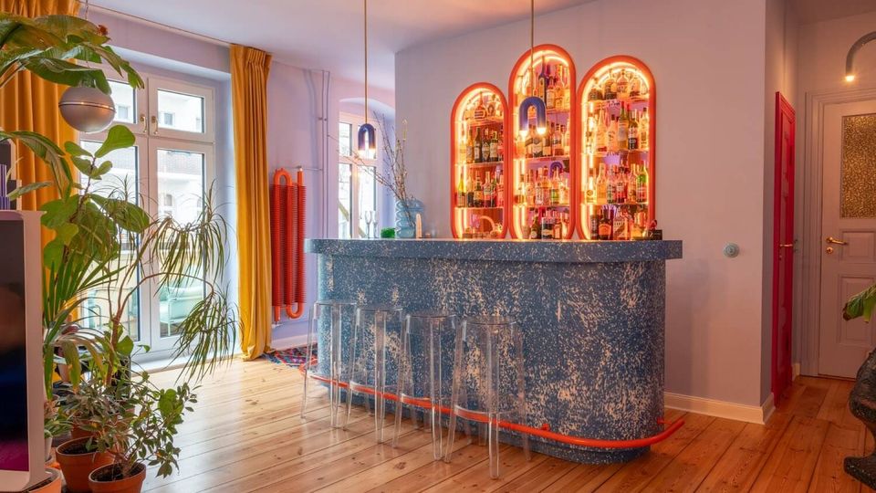 Bright, modern home bar area with a curved blue speckled counter, clear acrylic stools, and three arched backlit bottle shelves mounted on a pale wall. Large windows with mustard-yellow curtains, hanging pendant lights, indoor plants, and wood plank flooring define the space.