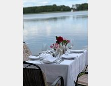 Ein gedeckter Tisch mit weißer Tischdecke, Weingläsern, Besteck und Blumenvasen, steht auf einer Terrasse mit Blick auf einen ruhigen See und bewaldetes Ufer im Hintergrund. Ein Segelboot ist in der Ferne auf dem Wasser sichtbar.