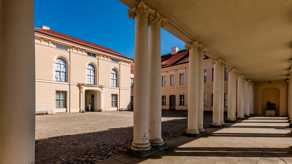 Neoclassical courtyard framed by a long covered colonnade with white columns and ornate capitals casting shadows on a stone walkway. Across the cobblestone square are pale stucco buildings with red tile roofs and tall arched windows under a clear blue sky.
