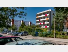 Mehrstöckige Wohngebäude mit Balkonen, die farbige Paneele aufweisen, umgeben von Bäumen und Sträuchern. Im Vordergrund befindet sich ein Parkplatz mit geparkten Autos und ein Fußweg.