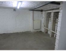 Kellerraum mit weißen verputzten Ziegelwänden, Betonboden und einer Decke mit sichtbaren Rohren und einer Leuchtstoffröhre. An der Wand befindet sich ein Heizkörper, und eine offene Mauerstruktur zeigt kleine Nischen.