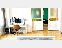 Arbeitszimmer mit modernem Schreibtisch, Bürostuhl und Computer vor einer weißen Wand. Im Hintergrund führen verzierte Schiebetüren mit milchigen und gelblichen Glaselementen in einen Raum mit einer grünen Wand und Esstischstühlen.