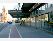 Eingangsbereich des Bezirksamts Steglitz in Berlin mit Glasfassaden und einem breiten Vordach; links ist eine Straße, daneben sieht man ein historisches rotes Backsteingebäude mit Turm.