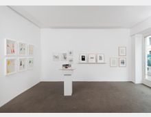 Ein minimalistisch gestalteter Galerieraum mit weißen Wänden, auf denen gerahmte Kunstwerke hängen, darunter abstrakte Zeichnungen und Grafiken. In der Mitte des Raumes steht ein weißer Sockel mit einem kleinen Modell oder Kunstobjekt; das Fenster auf der rechten Seite lässt Tageslicht herein.