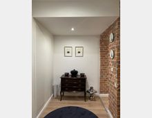 Ein kleiner Eingangsbereich mit einer Kommode aus dunklem Holz, darauf dekorative Gegenstände wie eine schwarze Vase. An der weißen Wand hängen zwei gerahmte Bilder mit Fischmotiven, rechts ist eine freiliegende Backsteinmauer mit aufgehängten Tellern sichtbar. Der Boden besteht aus hellem Holz, und ein runder, grauer Teppich liegt im Vordergrund.