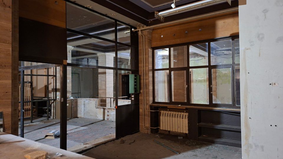Verlassener Innenraum eines ehemaligen Büro- oder Werkstattbereichs mit Glas- und Metalltrennwänden, hohen Fenstern und sichtbaren Deckenbalken. Abgeblätterte Wände, freiliegende Leitungen, ein Heizkörper unter dem Fenster und Bauschutt am Boden prägen die Umgebung.
