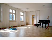 Heller Raum mit Holzboden, großen Fenstern mit weißen Vorhängen und einem Flügel auf der rechten Seite. In einer Ecke stehen ein Holzstuhl und Dekorationen, während Sonnenlicht durch die Fenster hereinfällt.