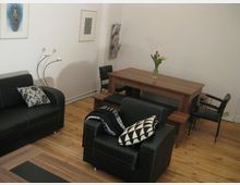 Wohnzimmer mit Holzboden, schwarzer Ledersitzgruppe, Holz-Esstisch mit passender Bank, modernen Dekokissen und einer Vase mit Blumen als Tischdekoration. Helle Wände mit dekorativen Bildern.