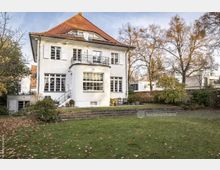 Weiße zweistöckige Villa mit rotem Walmdach, großen Fenstern und einem Balkon. Im Vordergrund befindet sich ein gepflegter Garten mit Rasen, Bäumen und herbstlichem Laub.