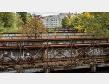 Verrostete Eisenbahnbrücke mit Graffiti und überwucherter Vegetation, im Hintergrund moderne Wohngebäude und zahlreiche Bäume.