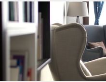 Wohnzimmeransicht mit einem beigefarbenen und einem blauen Sessel, einer Stehlampe mit hellem Lampenschirm im Hintergrund und einem Bücherregal am Bildrand. Im Hintergrund sind ein Fenster mit blauen Vorhängen und ein Kissen auf dem blauen Sessel sichtbar.