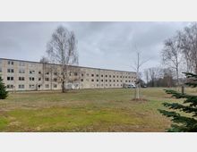 Ein schlichtes, vierstöckiges Wohngebäude mit rechteckigen Fenstern und heller Fassade steht in einer offenen Grünfläche. Im Vordergrund befinden sich kahle Bäume, ein kleiner Nadelbaum und ein geparkter weißer Transporter.