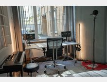 Ein schlichtes Musikproduktionsstudio in einem Raum mit großen Fenstern und Tageslicht. Zu sehen sind ein Schreibtisch mit Lautsprechern, ein Bürostuhl, ein Mikrofonständer, ein Keyboard mit Hocker und Kopfhörer, sowie graue Vorhänge und eine rote Teppichfläche.