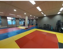 Ein Trainingsraum mit farbigen Matten in Rot, Gelb und Blau auf dem Boden; im Hintergrund stehen Fitnessgeräte, Hanteln und Boxsäcke. Große Fenster an einer Seite lassen Tageslicht in den Raum, während die Deckenbeleuchtung den Raum gleichmäßig erhellt.