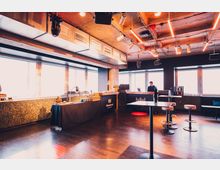 Industriell gestalteter Innenraum mit großen Fenstern, freiliegenden Deckenrohren und moderner Beleuchtung. Im Raum befinden sich mehrere Stehtische, eine Bar mit einem Mitarbeiter im Hintergrund und ein Buffet auf der linken Seite.