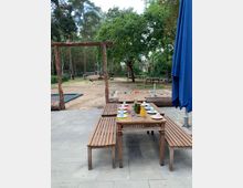 Holztische und -bänke stehen auf einer gepflasterten Terrasse, gedeckt mit bunten Bechern, Tellern und Kannen. Im Hintergrund ist ein Spielplatz mit Sandboden, Holzspielgeräten und Bäumen zu sehen.