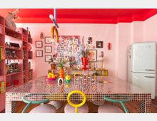 Bright, colorful kitchen-dining space with a glossy red ceiling, pink walls, and a large square mosaic-tiled table surrounded by modern chairs. Open shelving lines one wall, framed art and vases decorate the back wall, and a retro-style white refrigerator stands to the right.