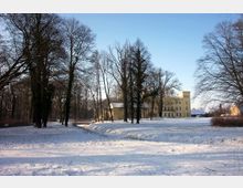 Schlosspark mit schneebedecktem Boden, hohen, kahlen Bäumen und einem gelben, historischen Gebäude im Hintergrund. Der Park enthält eine kleine Holzbrücke und ist von offenen Wiesen und bewaldeten Bereichen umgeben.