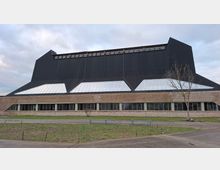 Ein modernes Gebäude mit einer schwarzen, steilen Dachstruktur, großen trapezförmigen Fenstern und einer unteren Fassade aus Holzverkleidung und Ziegeln. Im Vordergrund eine unbefestigte Wiese und ein Baum.