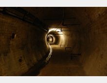 Ein unterirdischer, kurvenförmig verlaufender Tunnel mit Backsteinwänden, schwacher Beleuchtung und sichtbaren Rohren an der Decke. Der Boden ist teils feucht, und entlang der Wände befinden sich Metallhalterungen.