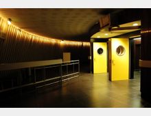 Ein halbdunkler Innenraum mit wellenförmigen Wänden und indirekter Beleuchtung; im Vordergrund gelbe Türen mit runden Fenstern, daneben ein Metallgeländer und ein dunkler Steinboden.