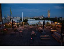 Dachterrasse bei Abenddämmerung mit Sitzbänken, Tischen und Pflanzen, dekoriert mit Lichterketten. Im Hintergrund ist eine städtische Skyline mit Industriegebäuden, Wohnhäusern und einem beleuchteten Hochhaus zu sehen.