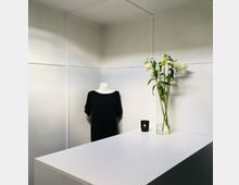 Eine minimalistische helle Innenraumszene mit einer weißen Wand, einer Schaufensterpuppe in einem schwarzen Kleid, einem weißen Tisch im Vordergrund, einer Vase mit weißen Lilien und einer schwarzen Kerze darauf.
