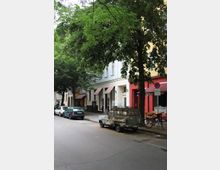 Straßenszene in einer Stadt mit mehrstöckigen Gebäuden, die helle Fassaden und Markisen über den Eingängen haben. Am Straßenrand stehen geparkte Fahrzeuge, darunter ein rostiges, älteres Auto, und Bäume säumen die Straße.