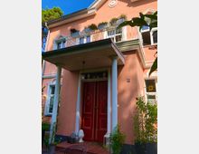 Rosa Hausfassade mit roten Doppeltüren und darüber einem kleinen Balkon mit bogenförmigem Geländer, geschmückt mit Blumenkästen. Das Gebäude hat weiße Fensterrahmen, einen überdachten Eingang mit zwei Säulen und eine Jahreszahl (1791) auf der Fassade.