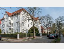 Eckgebäude in einer Wohngegend mit weißer Fassade und roten Ziegeldächern, umgeben von Bäumen und gepflegtem Zaun. An der Straße stehen geparkte Autos, und ein gepflasterter Gehweg verläuft entlang der Reihenhäuser.